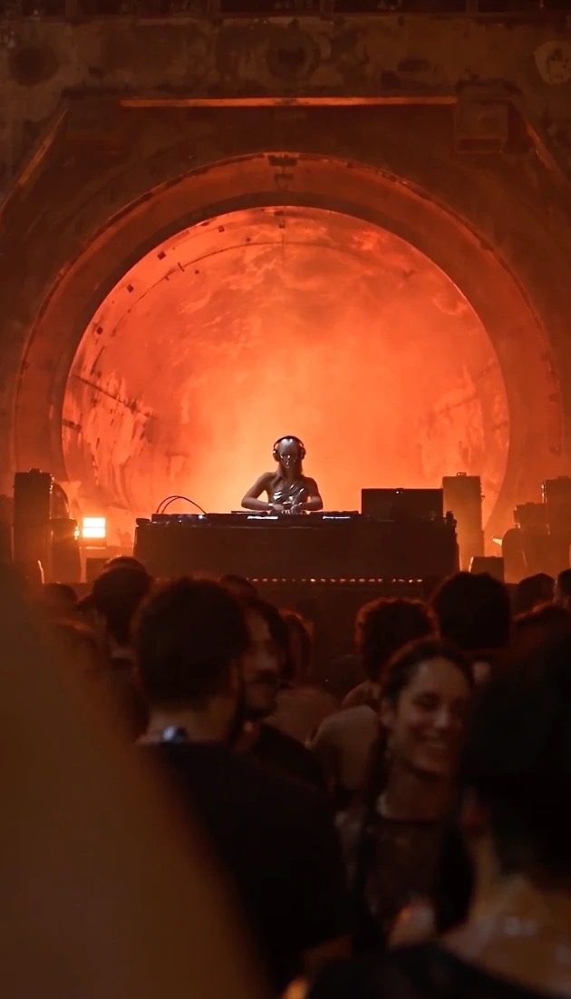 A masked DJ at the decks in front of a glowing red tunnel, a crowd in the foreground